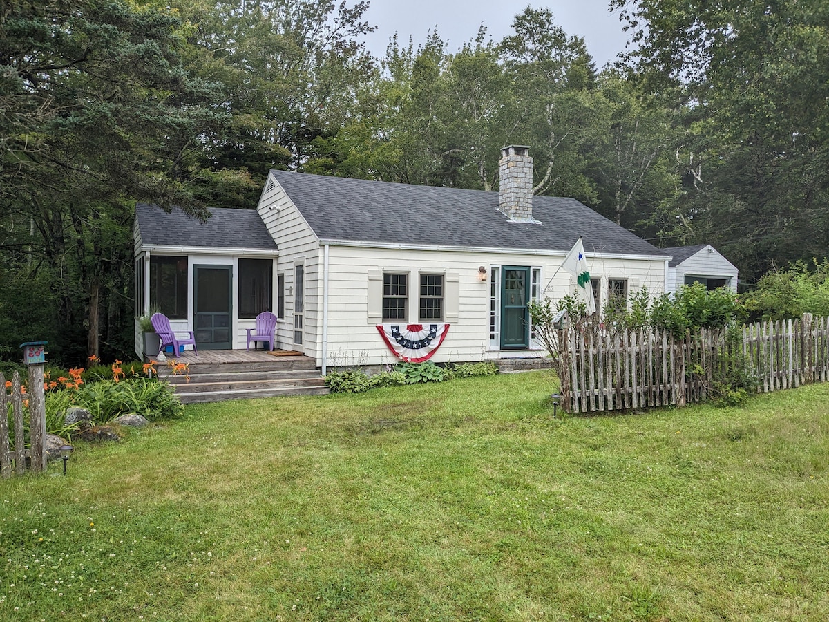 A charming cottage is framed by a well-maintained yard, featuring a wooden deck with purple chairs and seasonal decorations. A stone chimney is visible, and lush trees provide a natural backdrop. The cottage exudes a welcoming presence with white siding and a green door.