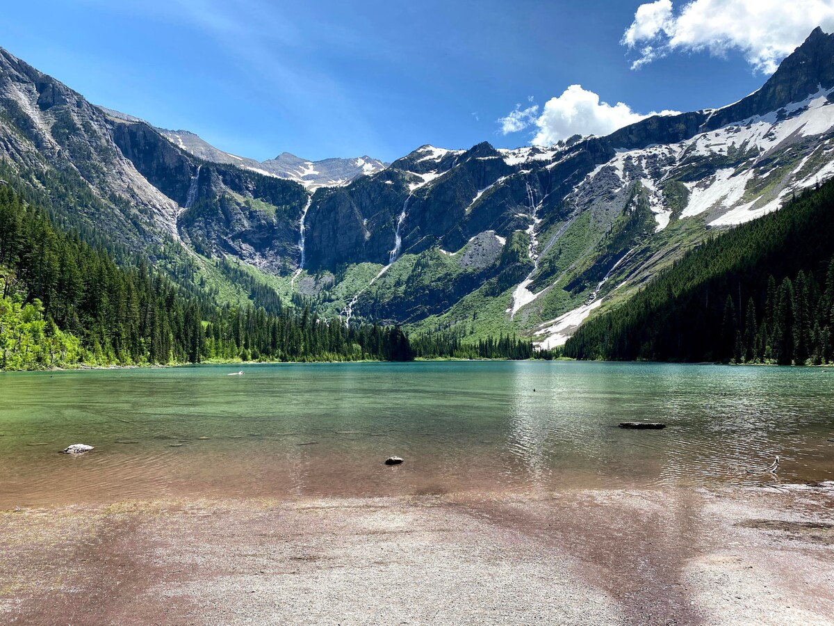 A serene mountainous landscape is reflected in clear turquoise waters. Lush green forests line the shore, harmonizing with snow-capped peaks in the background under a bright blue sky with scattered clouds.