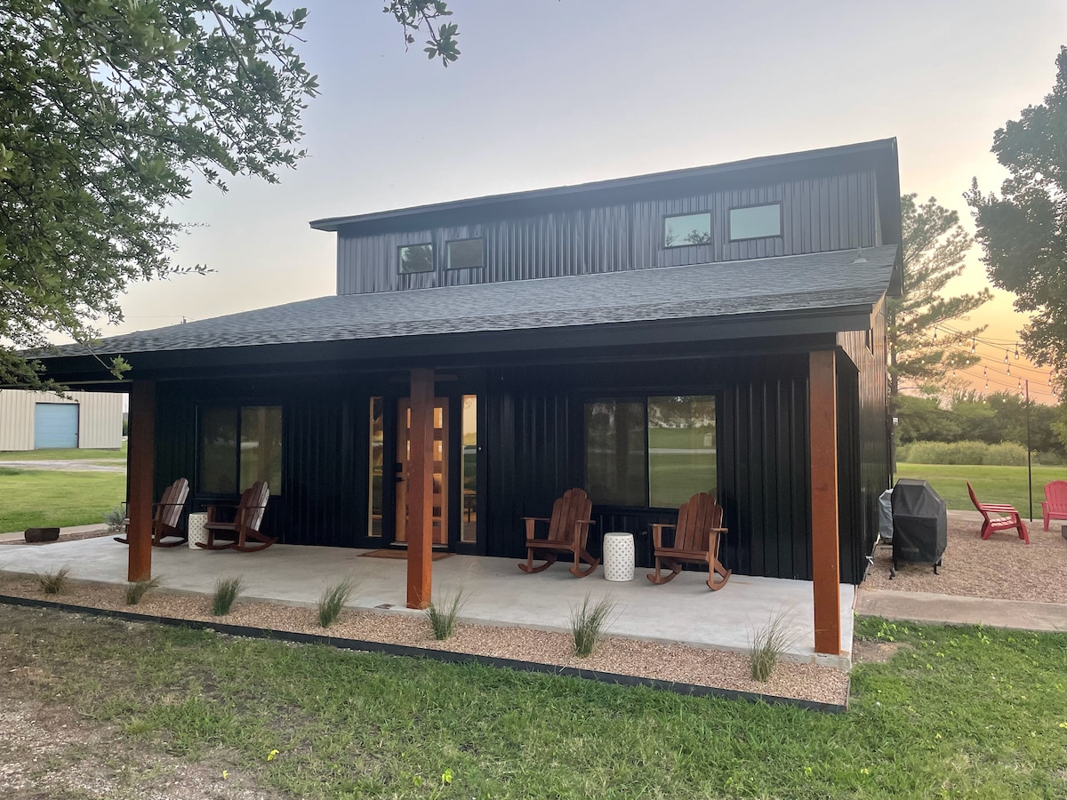 The exterior of a modern farmhouse is presented, featuring a wide porch with rocking chairs and a concrete patio. Large windows allow natural light to enter, while the building's black cladding contrasts with the surrounding greenery. A serene sunset is visible in the background.