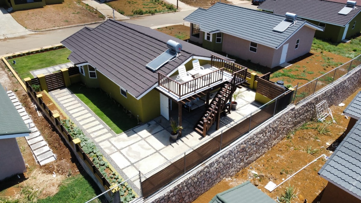 An aerial view showcases a yellow house with a terrace and seating area situated above a spacious patio. Surrounding areas include neatly maintained lawns and stone pathways, while nearby residences are visible in the background.