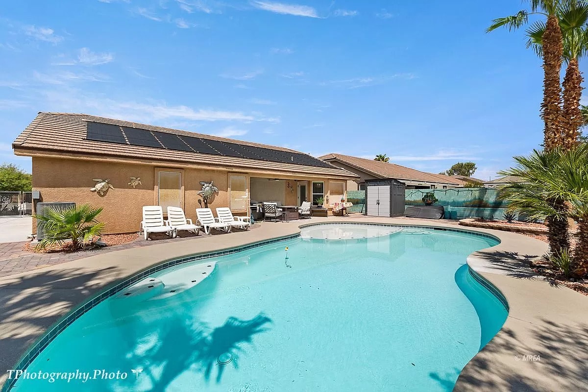 The inviting pool area features a spacious swimming pool surrounded by palm trees and lounge chairs. Bright sunlight illuminates the water, highlighting its clear blue reflections. A shaded patio area and landscaping elements create a relaxed outdoor environment.