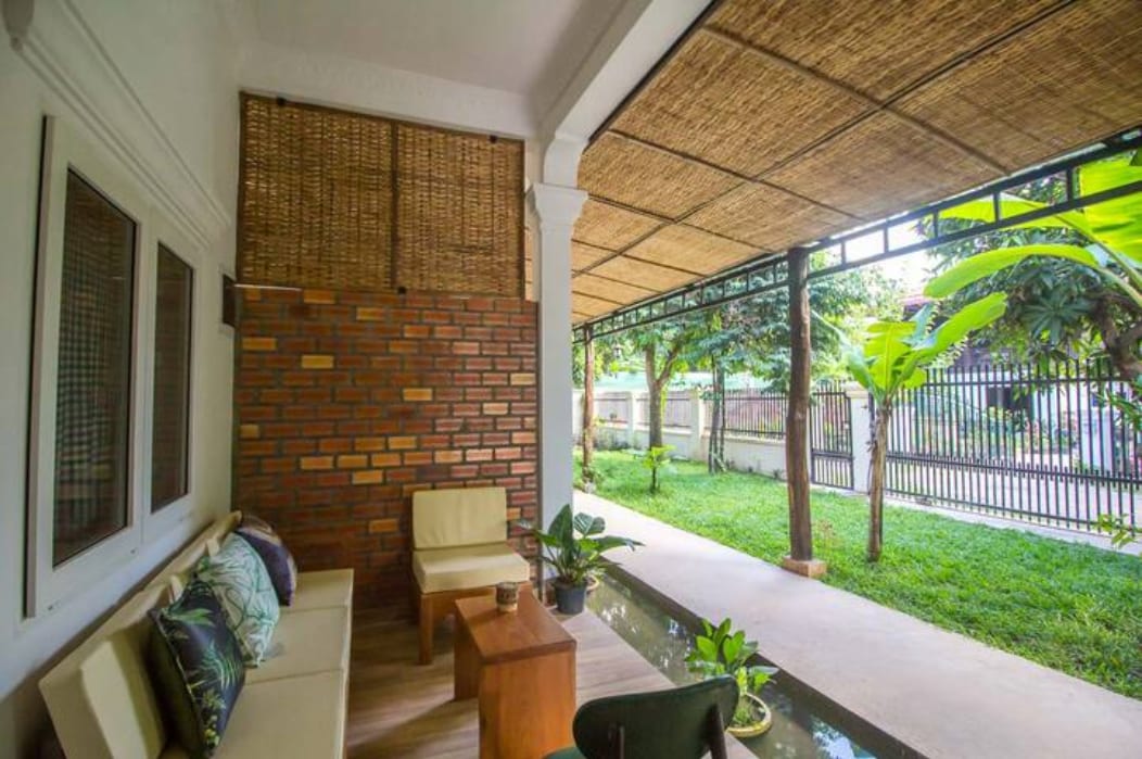A shaded outdoor seating area is featured, showcasing two light-colored chairs and a small wooden table. A lush green pathway is visible, bordered by tropical plants, with a textured brick wall adding warmth to the space.