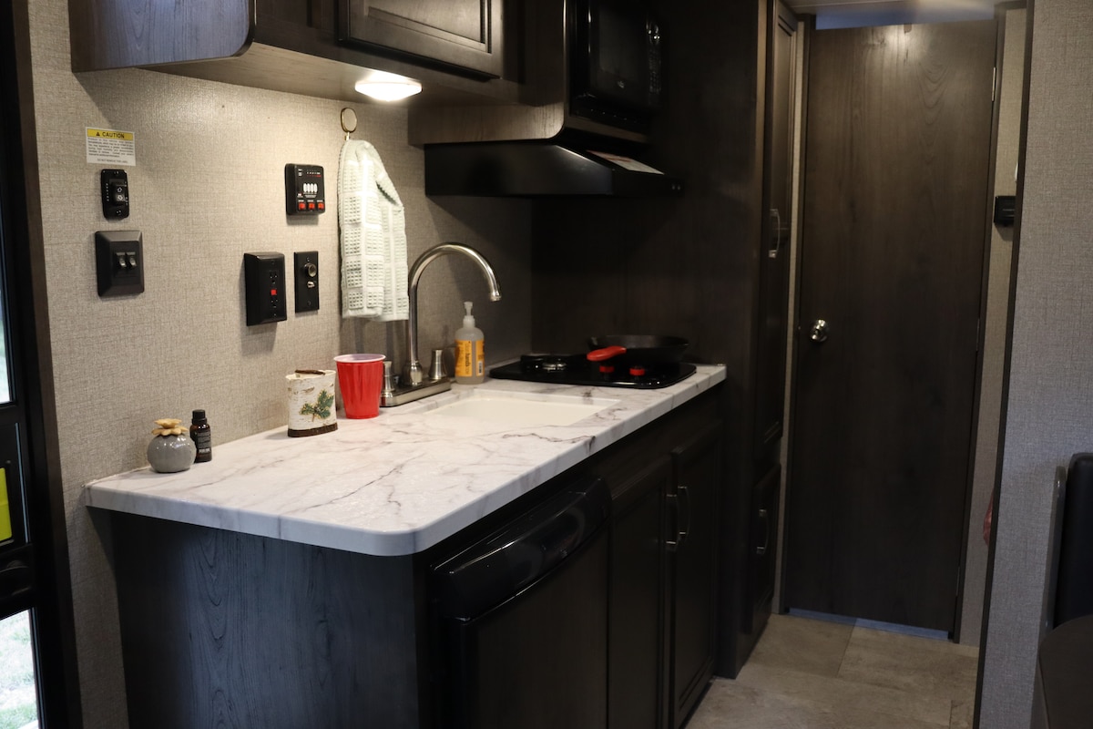 A compact kitchenette is displayed, featuring a marble countertop with a sink, stove, and microwave. Black cabinetry offers storage, while kitchen necessities are arranged neatly. The space is illuminated by overhead lighting, creating a functional cooking area adjacent to the entryway.