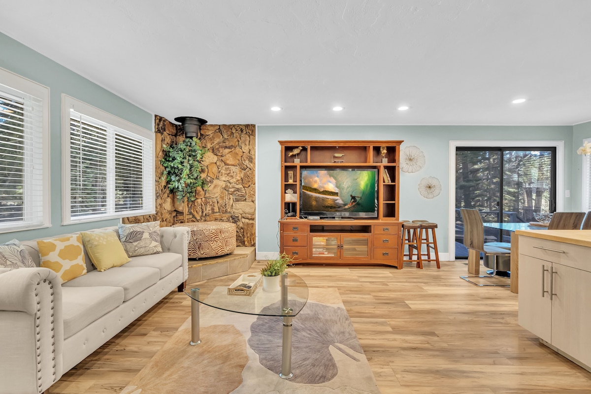 A welcoming living area features a light-colored sofa paired with colorful accent pillows. A large TV is mounted on a wooden console, complemented by a round glass coffee table. Natural light streams in through several windows, enhancing the cozy atmosphere.
