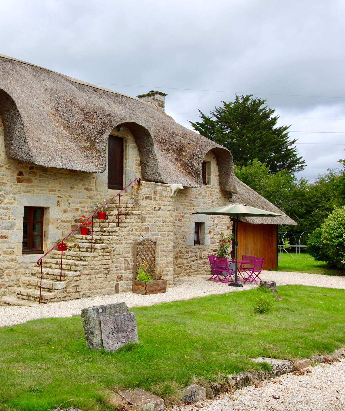 The exterior shows a traditional stone cottage with thatched roofing. A small stone staircase leads to the entrance. Two purple chairs and a table are positioned on a gravel pathway surrounded by green grass. Lush foliage forms a natural border on one side.
