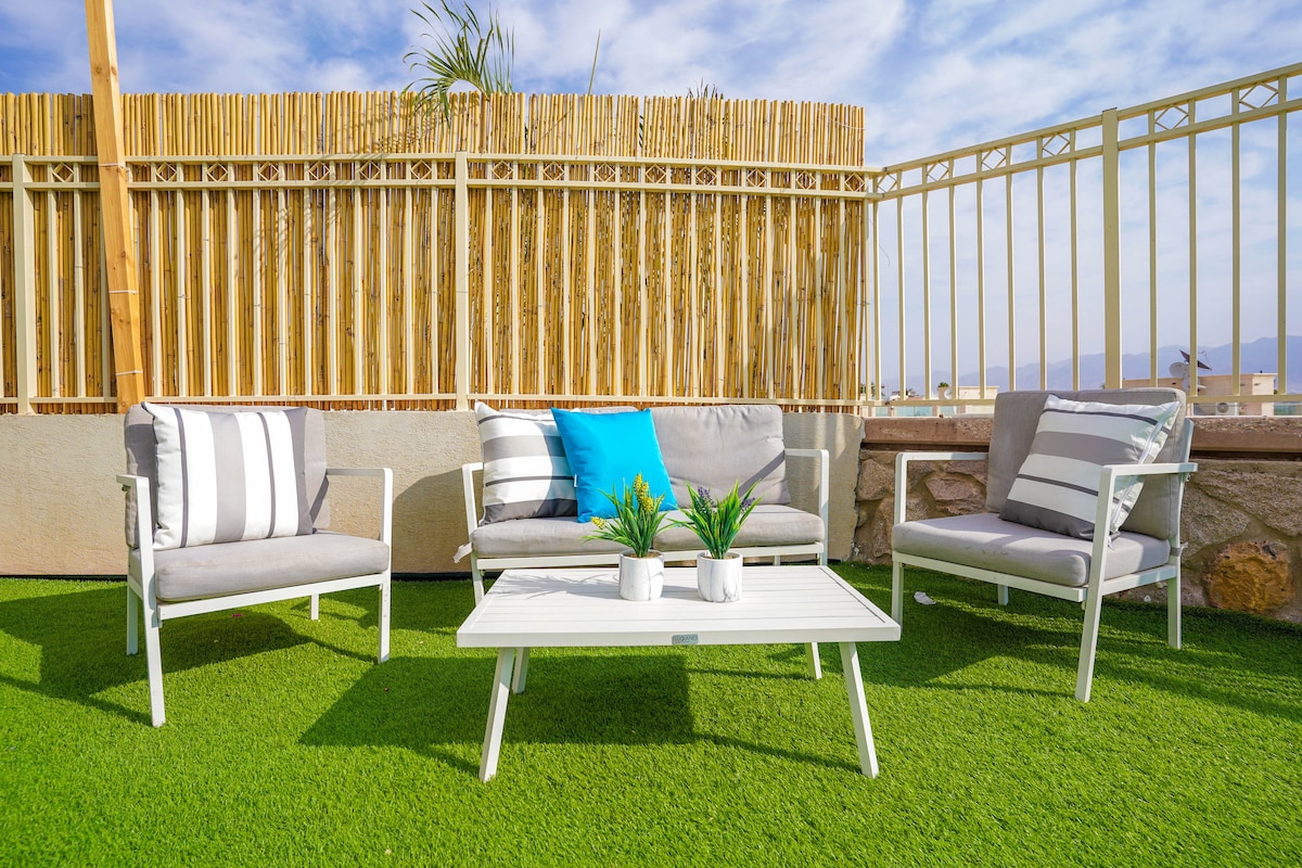 An outdoor seating area features a low table surrounded by four cushioned chairs, set on a patch of green grass. The space is enclosed by a bamboo fence, with light clouds visible in a blue sky above.