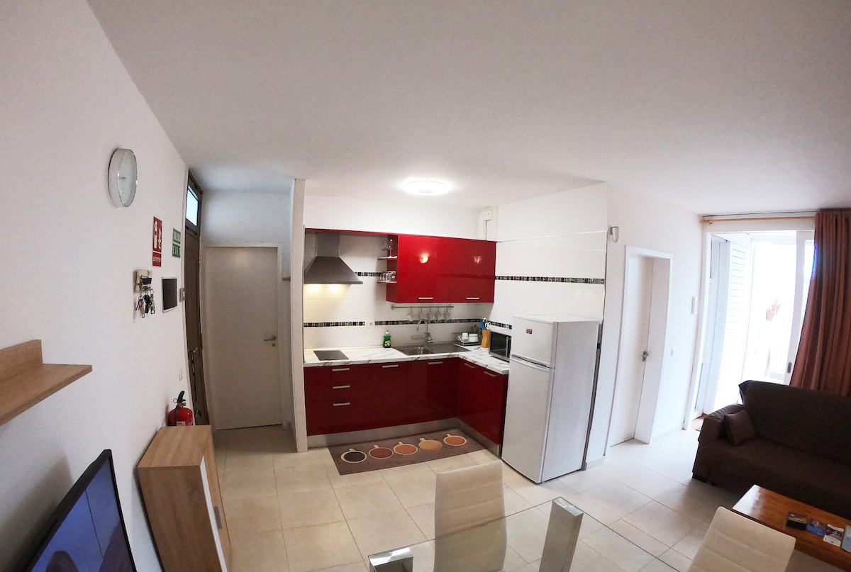 A modern kitchen area is showcased, featuring red cabinetry and stainless steel appliances. The space includes a rectangular dining table surrounded by six chairs. Natural light enters through a nearby window, illuminating the tiled floor and enhancing the sleek design.