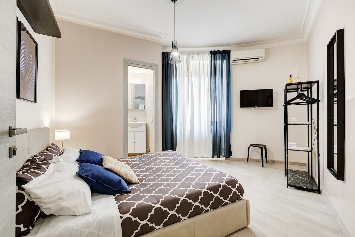 A spacious bedroom is featured with a large bed dressed in neutral tones, accented by blue pillows. Natural light floods the room through floor-to-ceiling curtains. A modern television is mounted on the wall, and a clothing rack is positioned in the corner, enhancing comfort and functionality.