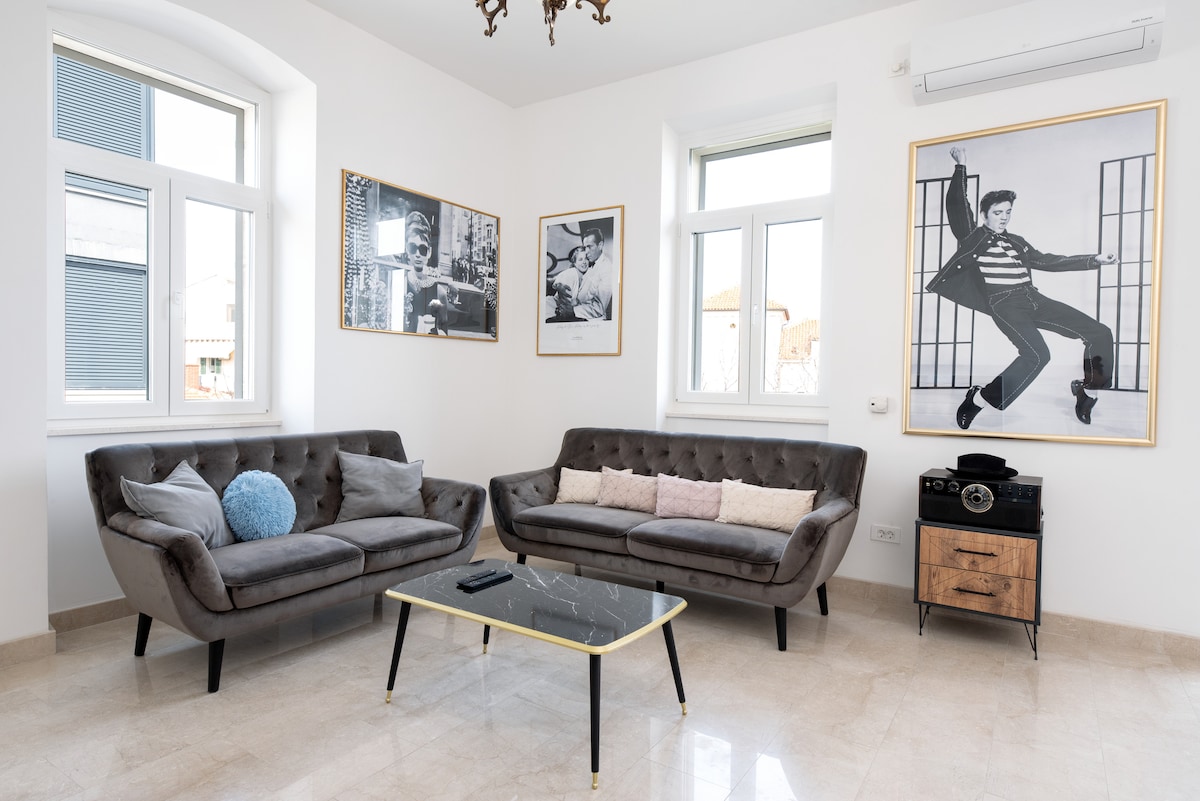A bright living area features two stylish sofas facing a glass coffee table. The walls are decorated with black-and-white photographs and a large framed artwork, enhancing the modern aesthetic. Natural light streams through large windows, creating an inviting space.