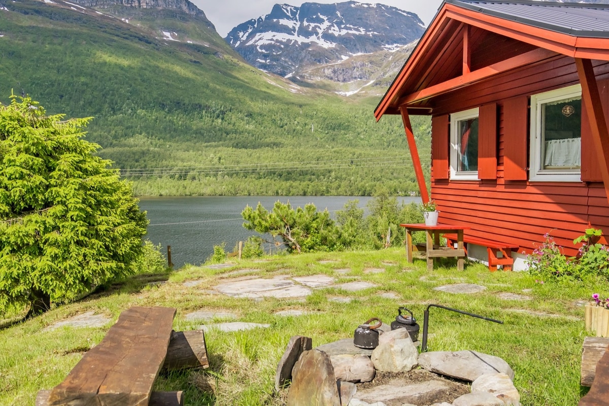 A cozy outdoor seating area offers a wooden picnic table and benches surrounding a stone fire pit. Lush green grass contrasts with the red cabin and is framed by views of the serene lake and majestic mountains in the background.
