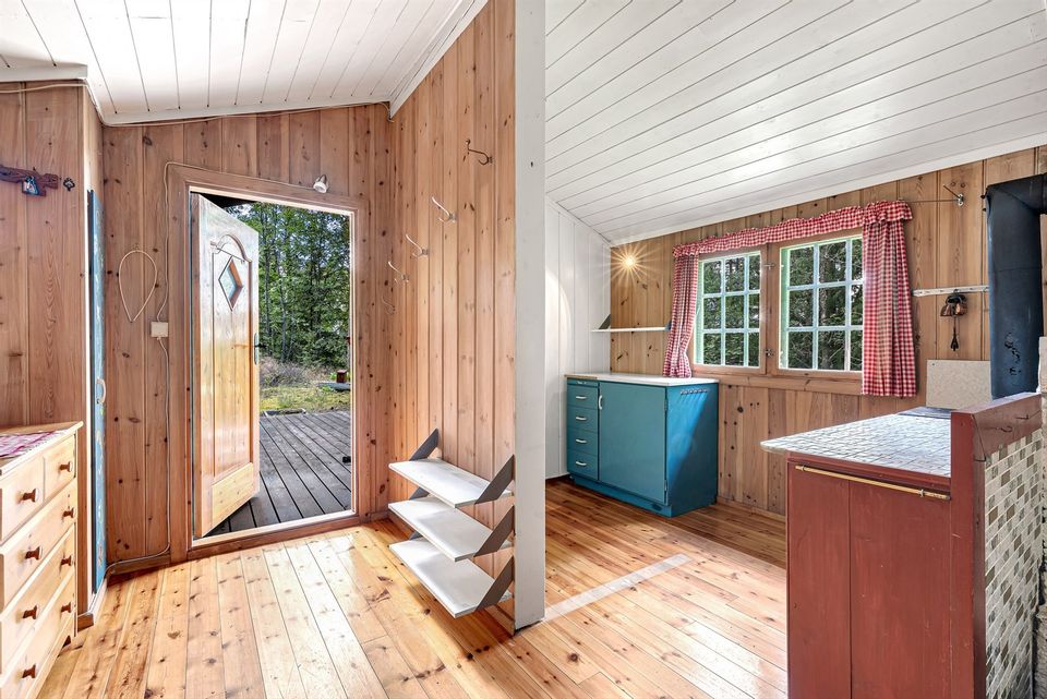 Wooden walls and a bright entry greet guests upon entering. A door with decorative glass opens to the outdoors, while a blue kitchenette is positioned beside a window adorned with checked curtains. Natural light illuminates the polished wooden flooring throughout the space.
