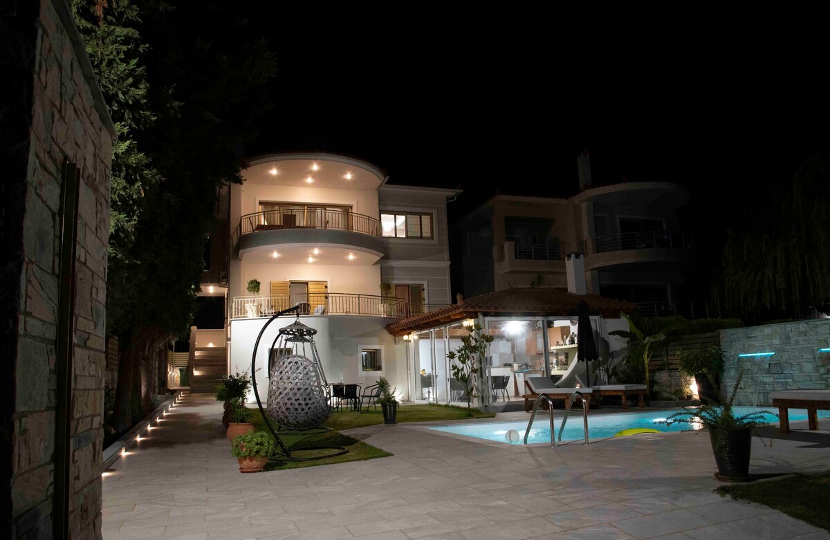 The villa is illuminated at night, showcasing its elegant architecture. A spacious outdoor area is visible, featuring a pool surrounded by lounge chairs. Soft lighting highlights the greenery and pathways, while a covered barbecue area is present nearby, integrating indoor and outdoor living.