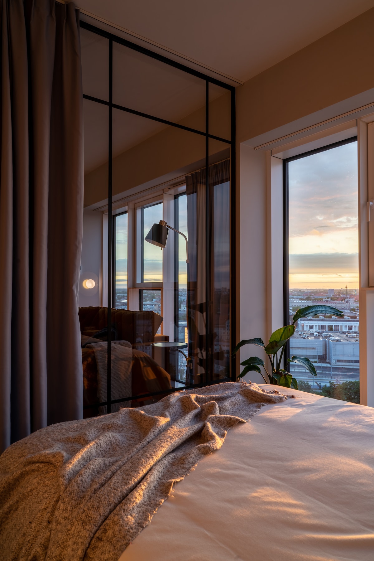 The bedroom features a spacious bed with a soft blanket draped across it. A large window offers a view of a sunset over the city. A sleek mirror reflects the light from a nearby lamp, while a green plant adds a touch of nature to the space.