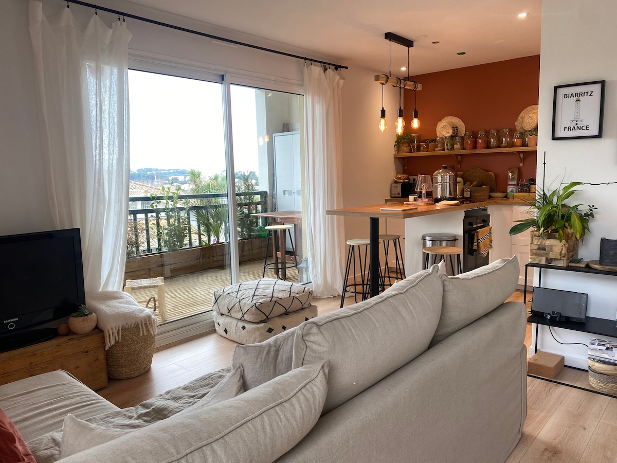 A bright living space features large sliding glass doors that open to a terrace, allowing for plenty of natural light. A cozy sofa and a pouf complement the contemporary decor. The kitchen island is equipped with bar stools, and decorative items enhance the ambiance.