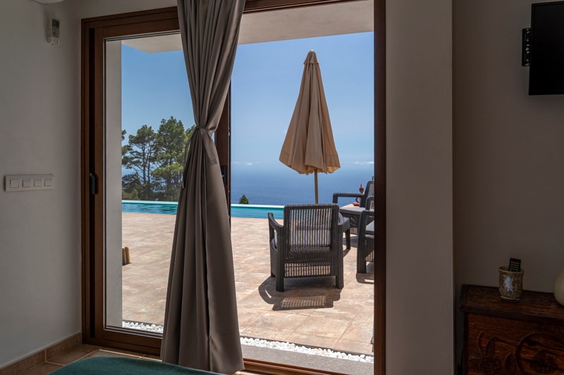 A large glass sliding door frames a view of an outdoor seating area, featuring a table and umbrella overlooking the infinity pool. Natural light fills the space, highlighting the simple decor of the interior and the tranquility of the setting.
