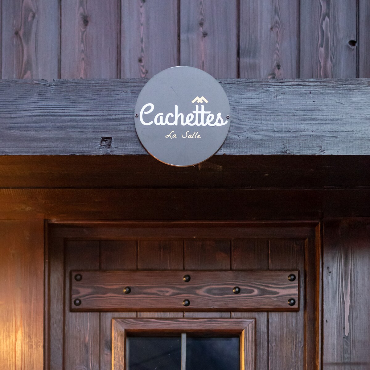 A round sign is displayed on a wooden wall, featuring the text 'Cachettes' with the location 'La Salle' below. The rustic wooden door beneath the sign is framed by dark wood, providing an inviting entrance.