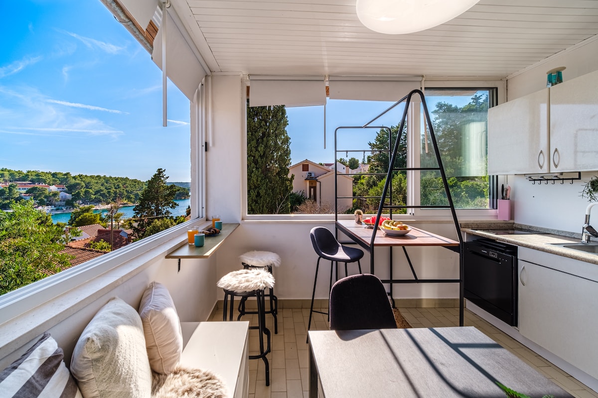 A bright and airy kitchen area features large windows providing a view of the surrounding nature and bay. Modern furnishings include a dining table with chairs and a small sofa. A breakfast bar adds additional seating, enhancing the functional layout of the space.