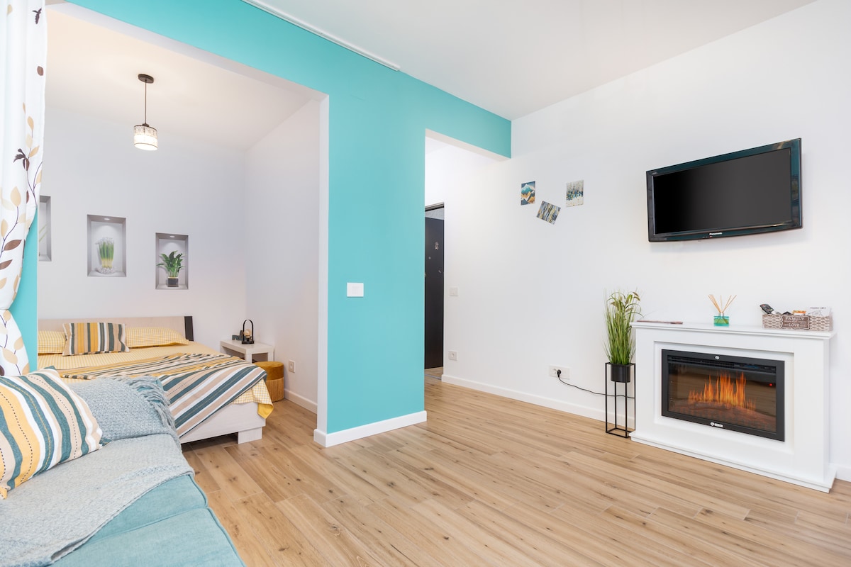 A bright living area features a cozy seating arrangement and a modern electric fireplace. A spacious bed with a yellow duvet is visible in the adjoining sleeping area. Decor includes framed botanical prints and a wall-mounted TV, enhancing the relaxed atmosphere.