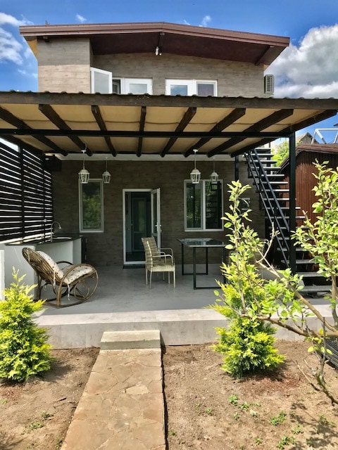 An exterior view of a two-story cottage features a spacious patio with outdoor seating, including two wicker chairs and a small table. A pathway leads to the entrance, framed by vibrant greenery and climbing plants. Large windows and a staircase add visual interest to the facade.