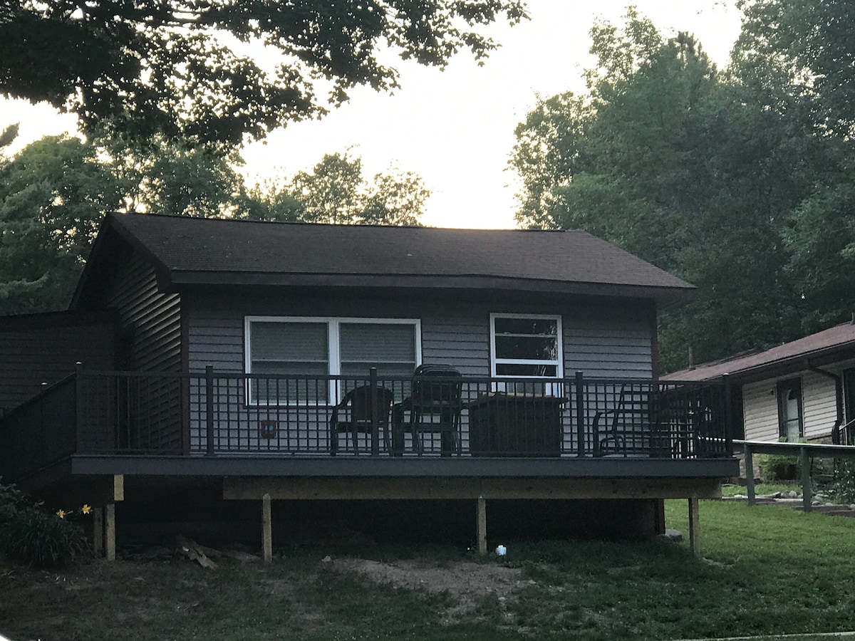 A cabin is depicted, featuring a spacious front deck equipped with several black chairs and a small table. The structure has a classic design with a brown roof and multiple windows, set amidst a backdrop of greenery.