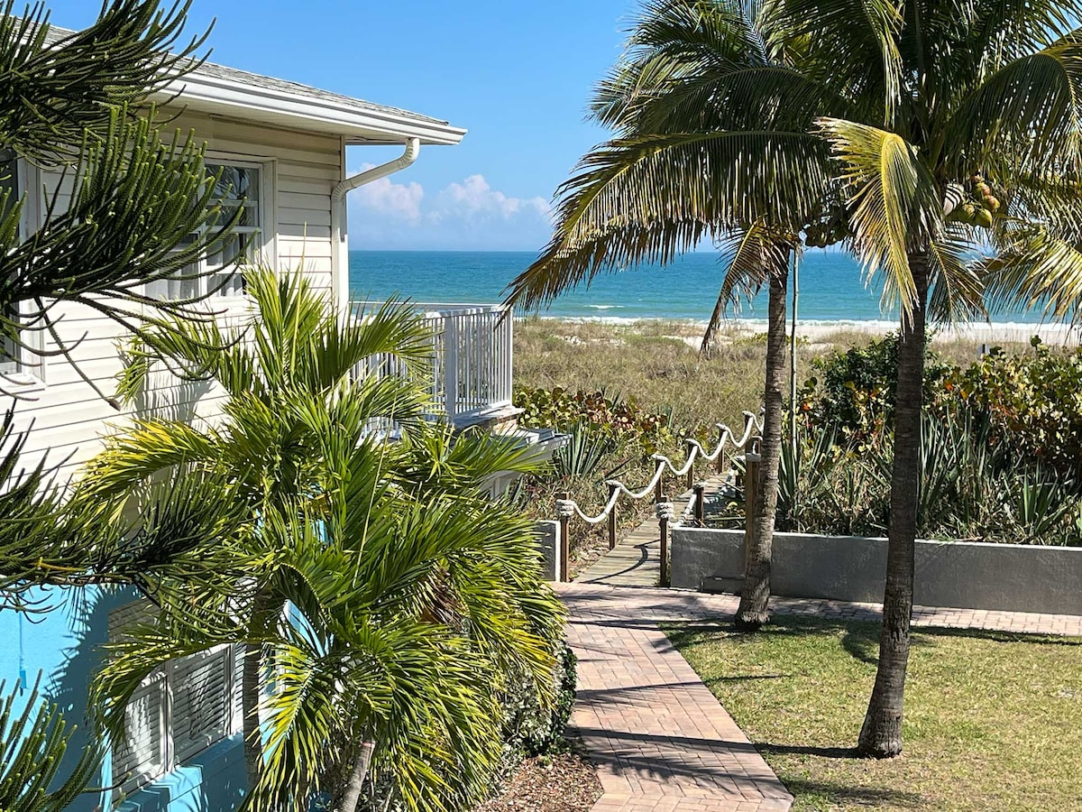 Lush tropical greenery frames the view of the oceanfront, creating a serene backdrop. The blue water glistens under the clear sky, with soft clouds visible in the distance. The pathway leads from the property to the private beach, enhancing the inviting coastal atmosphere.