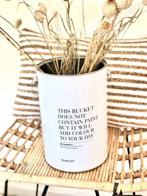 A decorative bucket filled with dried grasses is positioned on a woven chair. A white and black text printed on the bucket adds a modern touch. Striped cushions in neutral tones provide a soft contrast to the natural elements.
