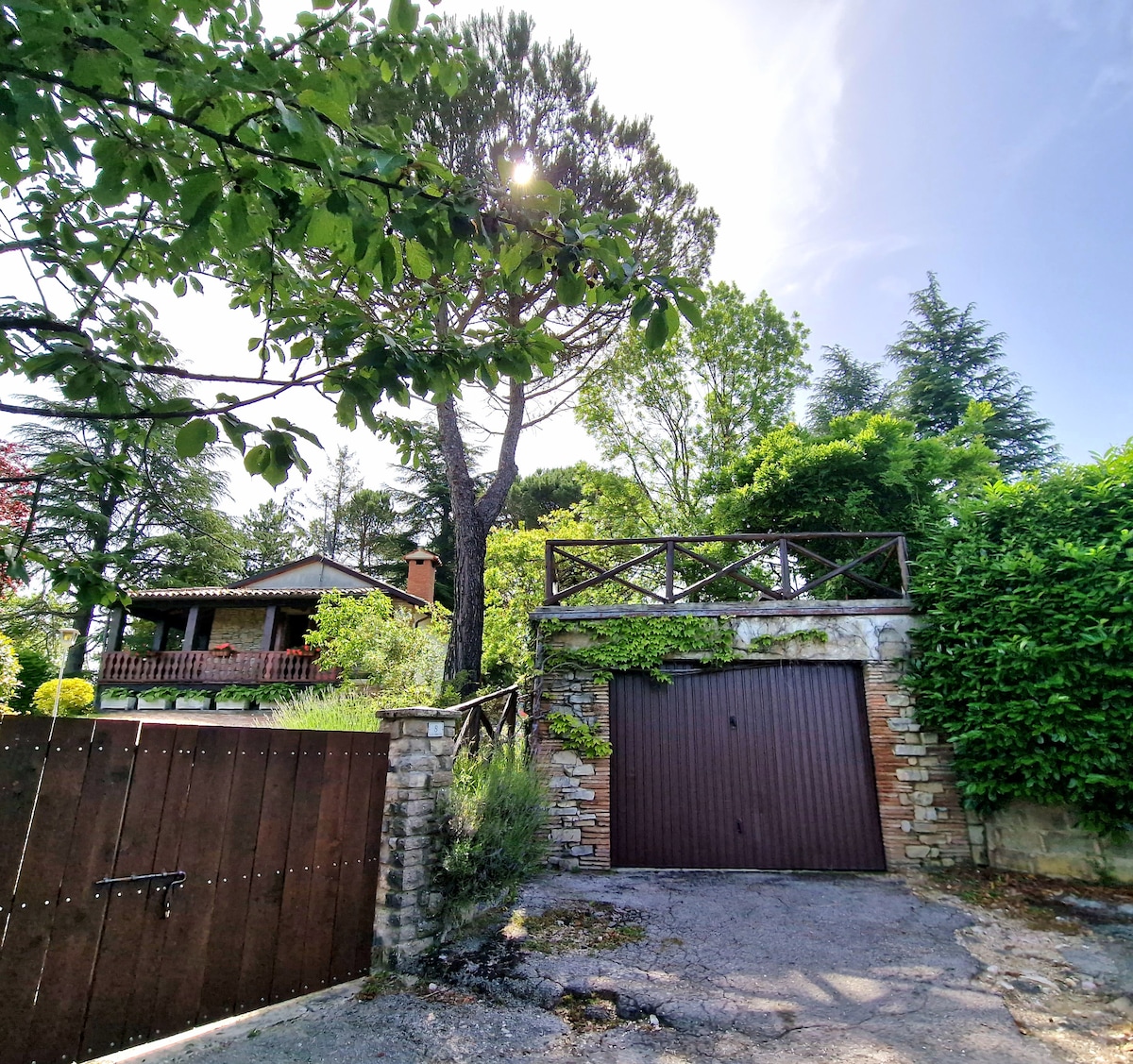 A serene entrance to the villa is framed by lush greenery, featuring a rustic wooden gate and a stone wall. The driveway leads up to a securely closed garage, surrounded by trees and vibrant foliage, under a clear blue sky.