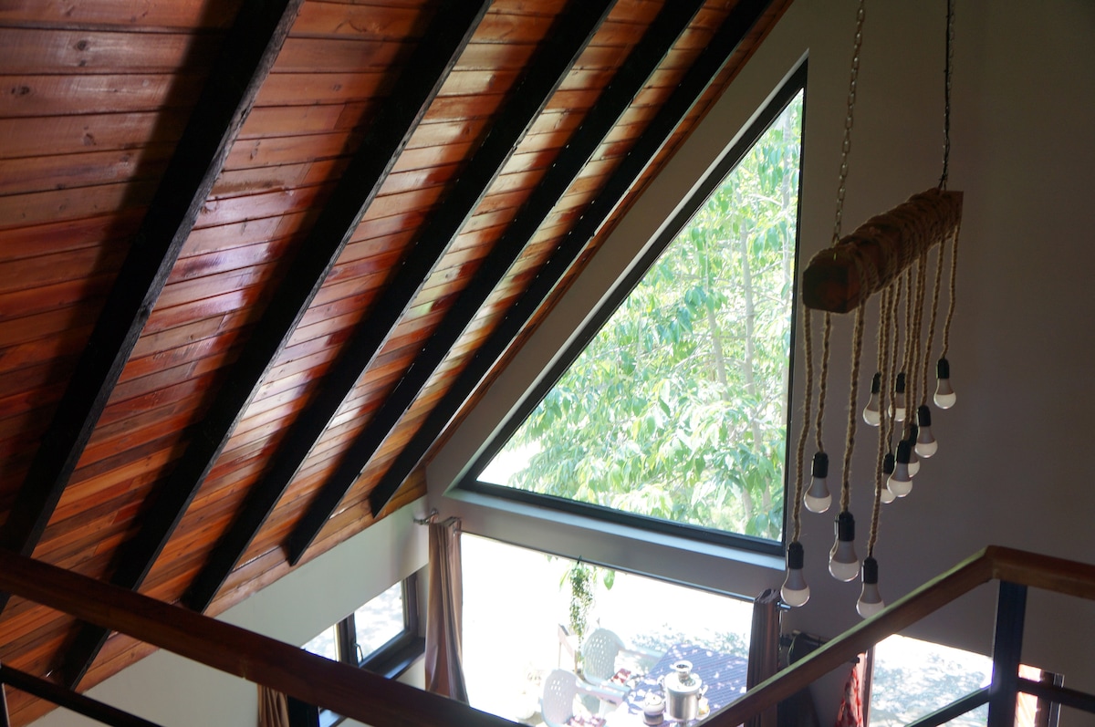A high ceiling with a sloped wooden structure is highlighted, featuring large triangular windows that allow natural light to fill the space. A collection of hanging lights can be seen, adding a modern touch to the rustic decor. Greenery is visible outside through the window.
