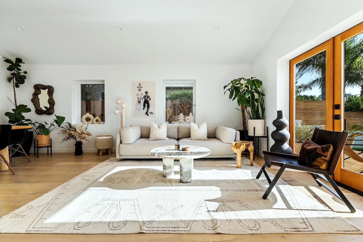A bright living area features a spacious sofa adorned with soft cushions, complemented by a round coffee table with a textured surface. A light-colored area rug covers the wooden floor. Natural light filters through large windows, adding warmth to the greenery arranged within the space.