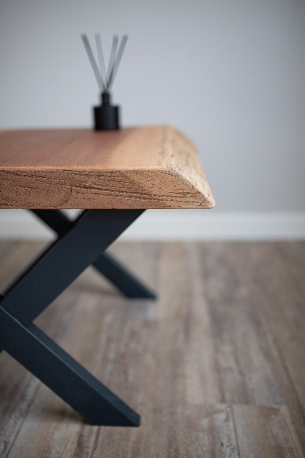 A wooden table with a natural edge is set atop a dark, cross-legged base, presenting a modern design. A black diffuser with reed sticks stands prominently on the table, enhancing the minimalist aesthetic of the space.