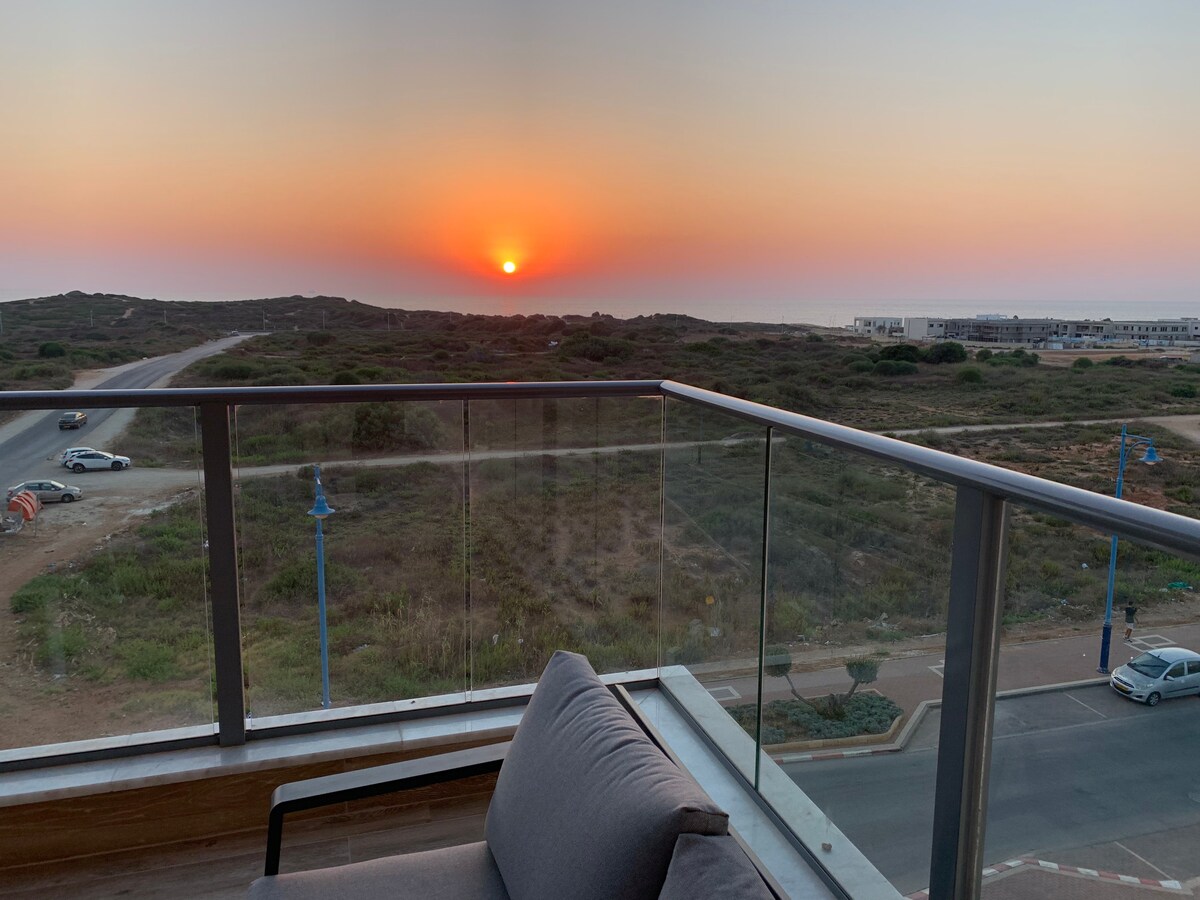 A balcony offers a view of the sunset over the horizon, with soft hues of orange and purple in the sky. A comfortable seating area is positioned on the balcony, providing a serene place to enjoy the outdoor scenery.