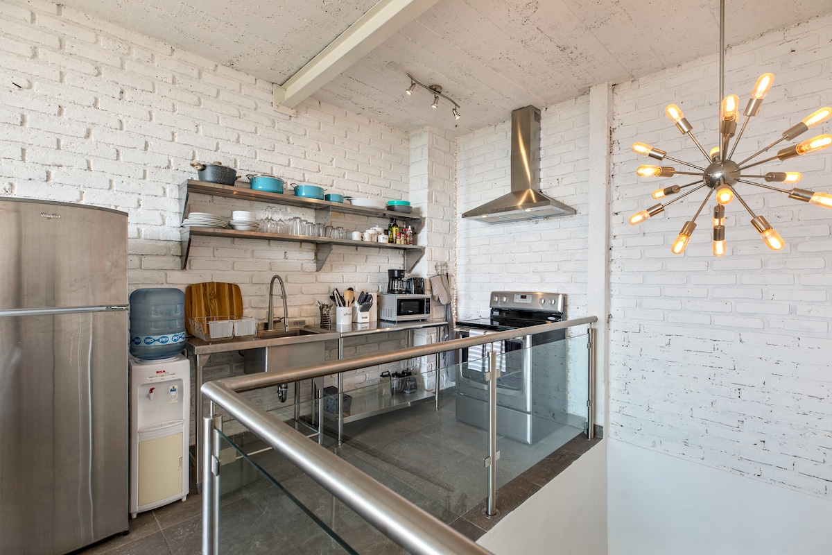 A modern kitchen is highlighted with stainless steel appliances and open shelving. A refrigerator stands alongside a sleek stove, while a variety of cookware and utensils are neatly arranged on the shelves. Light from a unique chandelier enhances the airy, minimalist design.