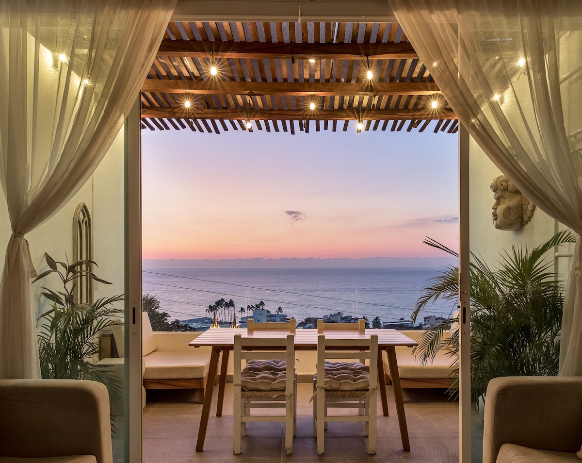 A dining area is positioned on a balcony with ocean views, featuring a wooden table surrounded by chairs. Strings of warm lights create a cozy ambiance, and the soft glow of sunset is visible over the water, enhancing the tranquil setting.
