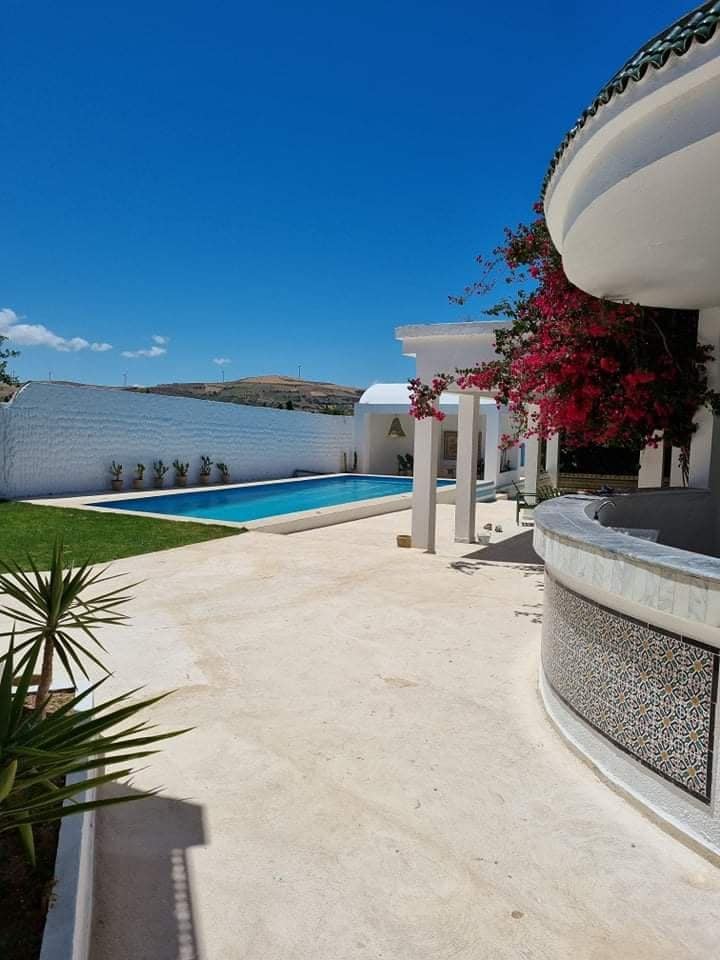An outdoor area is displayed, featuring a private pool surrounded by lush greenery. Vibrant flowers adorn the white walls of the villa, while sunlit tiles lead to a shaded terrace. Clear blue skies add to the serene ambiance of this tranquil space.