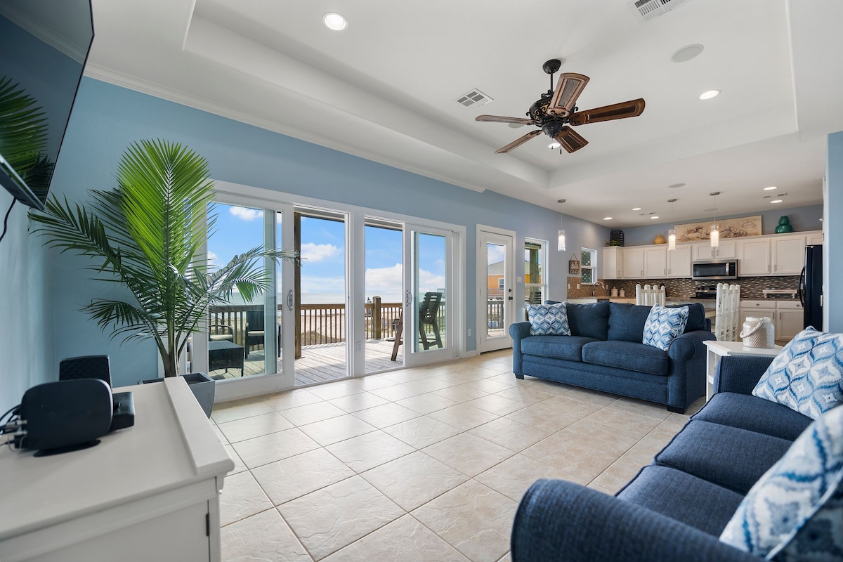 A spacious living area features a blue couch and armchair, with decorative cushions adding a pop of color. Large sliding glass doors open to a private balcony, allowing natural light to fill the space. A ceiling fan hangs above, complemented by light-colored tile flooring.