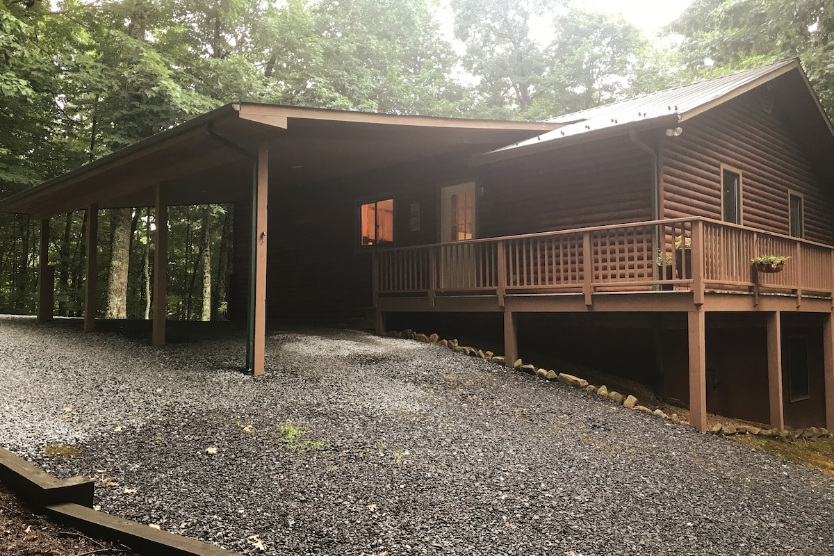 A wooden cabin is set amidst a wooded landscape, featuring a spacious covered porch that wraps around the front. A gravel driveway leads up to the entrance, while large windows allow glimpses of the warm interior. The surrounding trees create a serene, natural environment.