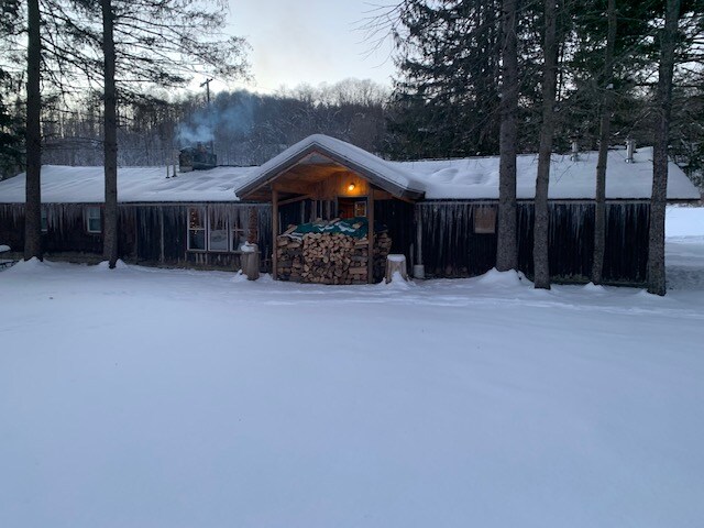 A log cabin, built with vertical logs, is nestled in a snowy landscape. The front porch features stacked firewood, and icicles hang from the roof. Pine trees surround the cabin, creating a serene winter setting.