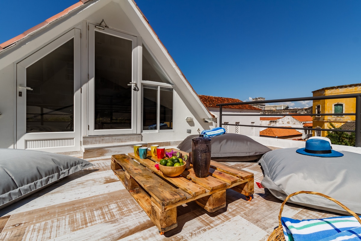 A terrace space features a wooden pallet table with fresh fruits and beverages. Soft gray cushions are arranged on the floor, providing comfortable seating. Large glass doors lead into the interior, with a clear blue sky overhead and views of nearby rooftops and buildings.