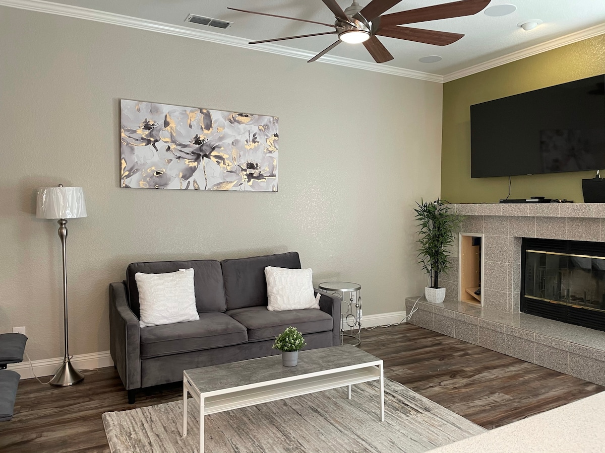 A comfortable living room features a gray sofa with white cushions, complemented by a light rug and a coffee table. A decorative painting hangs above the sofa, and a flat-screen TV is mounted on the wall above a polished fireplace. Indoor plants add a touch of greenery.