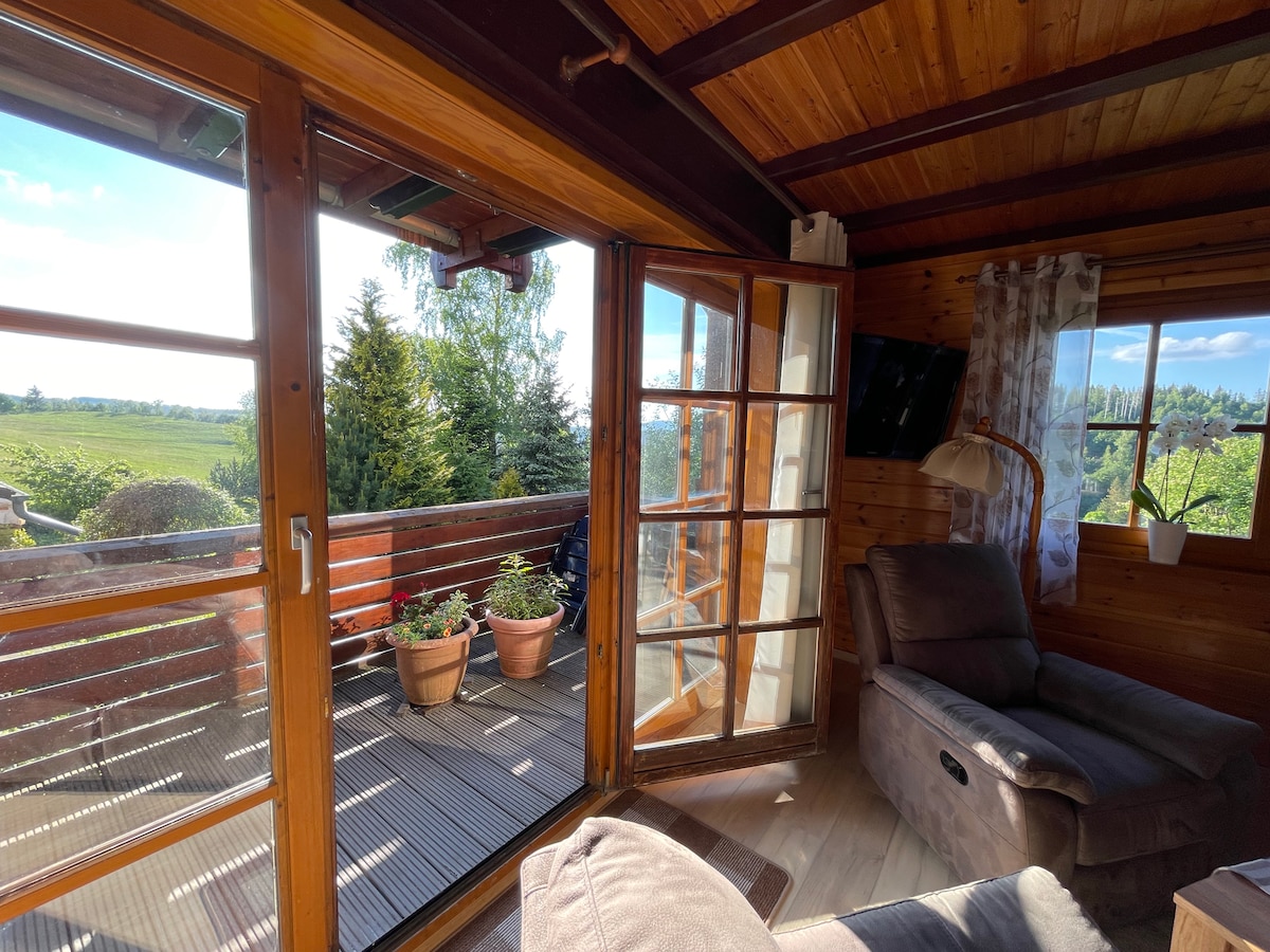 A wooden interior features large glass doors that open to a spacious balcony with potted plants. Natural light fills the room, highlighting the cozy seating area with a comfortable sofa. Scenic views of greenery can be observed from both inside and outside.
