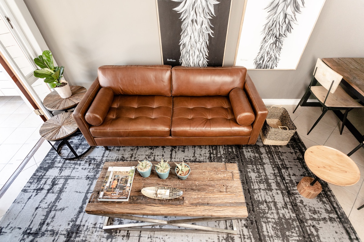 A comfortable leather sofa is centrally positioned within the living area, accompanied by wooden side tables. A textured rug enhances the space, while a wooden coffee table showcases a selection of magazines and decorative plants, creating a relaxed and inviting environment.