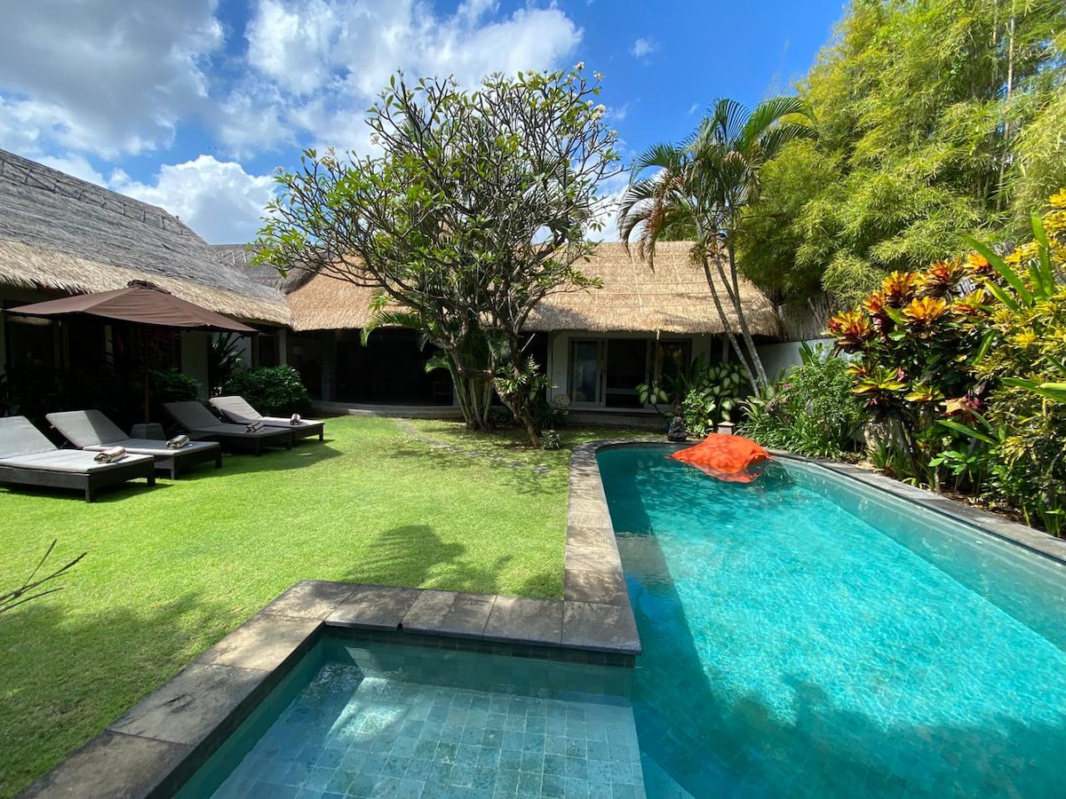 A private swimming pool is surrounded by a lush garden featuring varied tropical plants. Two sun loungers are positioned beside the pool, inviting relaxation. The thatched-roof building offers a view of the outdoor space, contributing to a serene atmosphere.