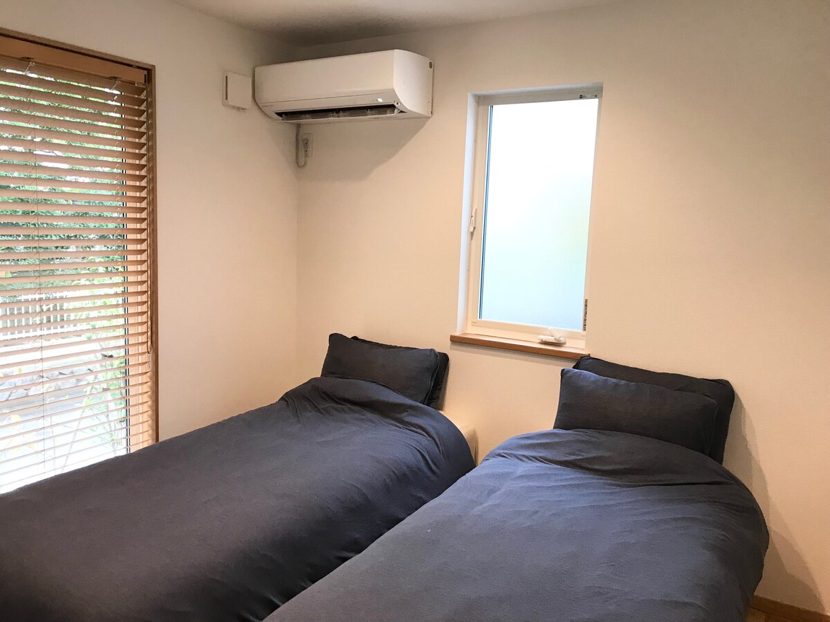 The bedroom features two single beds dressed in dark linens, positioned side by side. A window with blinds allows natural light to brighten the space. An air conditioning unit is installed on the wall, providing climate control.