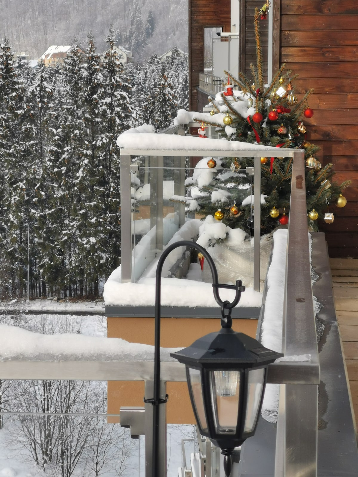 A balcony is featured with a snow-covered railing and a decorated Christmas tree adorned with red and gold ornaments. A classic lantern stands nearby, providing ambiance against a backdrop of snow-covered trees.