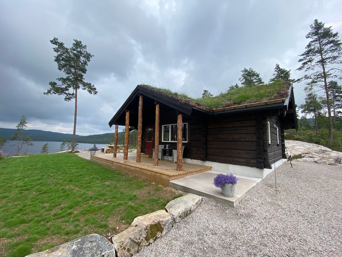 A traditional log cabin is set against a backdrop of trees and a lake. The structure features a green grass roof and wooden posts supporting the covered porch. A gravel area leads to a landscaped space with potted purple flowers, providing a serene outdoor setting.