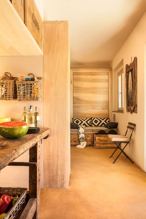 An open space is displayed, featuring a rustic wooden countertop and a small table with a chair. A cozy seating area with patterned cushions is visible in the background, complemented by a natural wood wall. Light enters through a nearby window, creating a warm atmosphere.