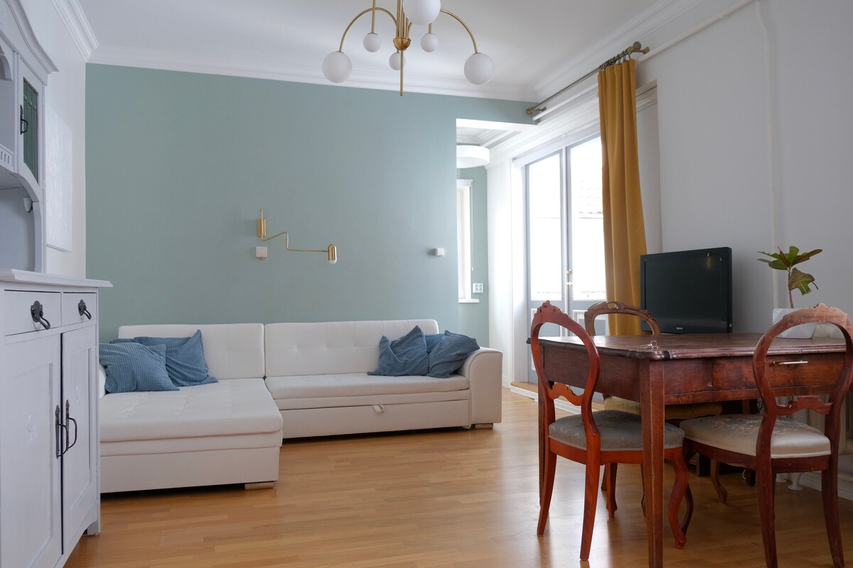 A bright living area features a white sofa positioned against a soft green wall. A wooden dining table is accompanied by four chairs, while a television stands nearby. Natural light enters through large windows, and warm curtains frame the space, enhancing its welcoming atmosphere.
