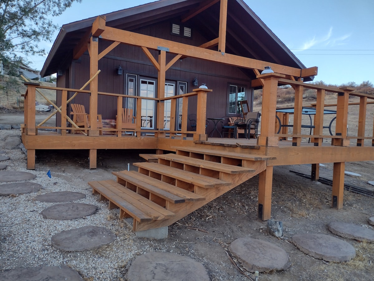 The exterior of a rustic glamping cabin is showcased, featuring a spacious deck with wooden steps leading to a welcoming entrance. Chairs and a table are positioned on the deck, with natural stone pathways leading through the surrounding landscape.