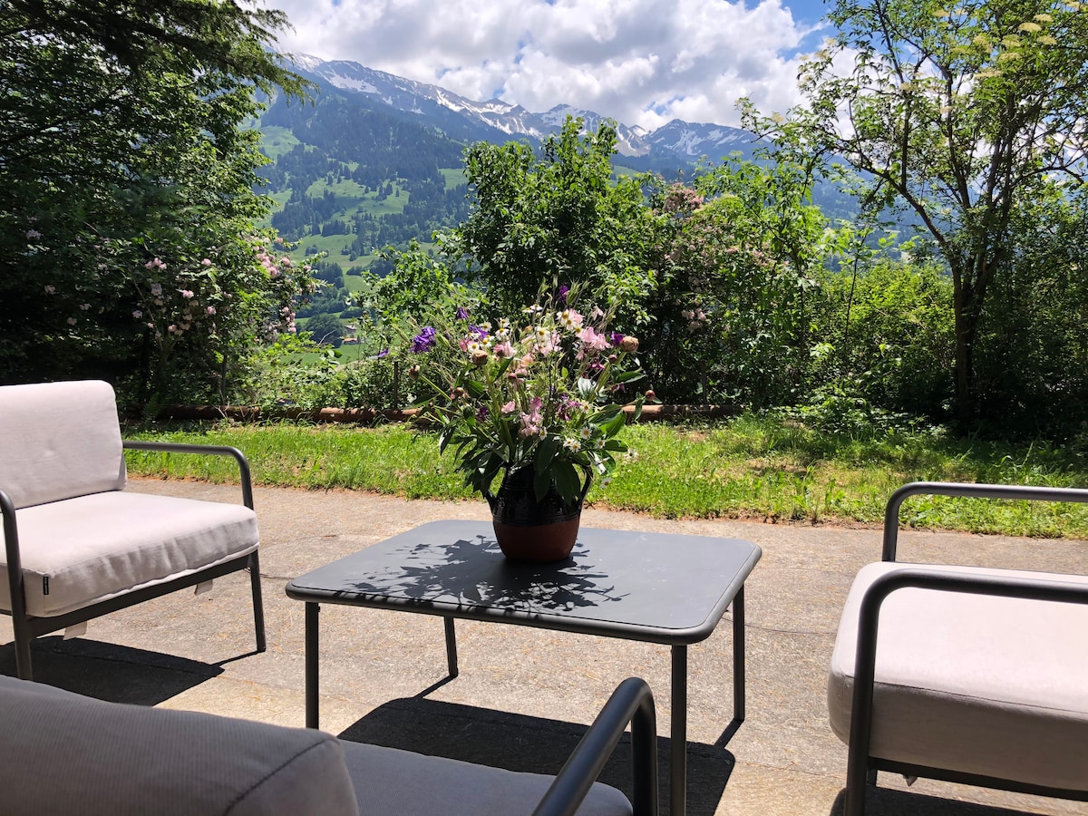 A private outdoor area features a table surrounded by four comfortable chairs. A potted flower arrangement adds color to the setting, with expansive mountain views in the background. Lush greenery contributes to a tranquil atmosphere, ideal for enjoying coffee or sunshine.