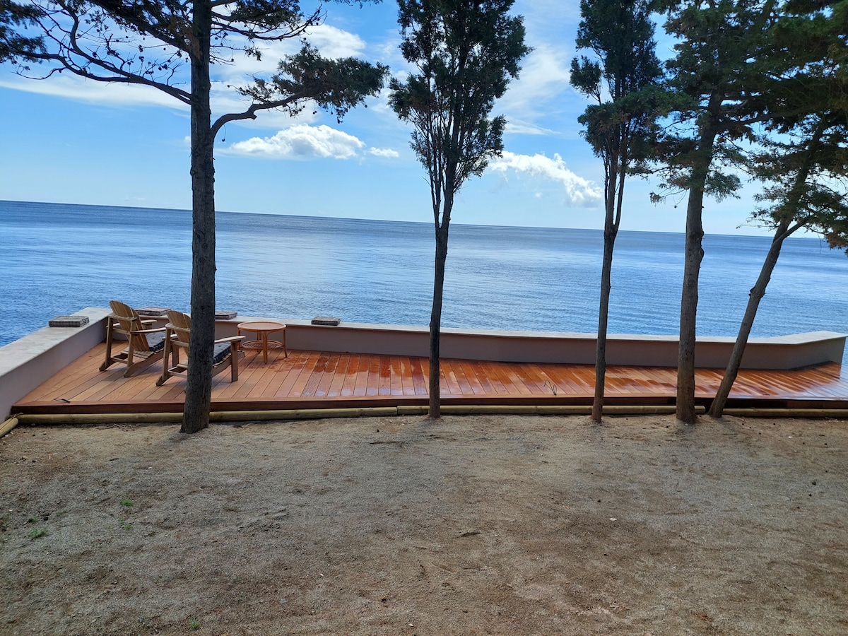 A wooden terrace offers direct views of the sea, framed by three tall trees. The spacious area includes two wooden chairs, inviting relaxation while enjoying the coastal scenery. Calm waters and a clear sky create a serene environment.